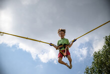 The Boy Is Jumping On A Bungee Trampoline. A Child With Insurance And Stretchable Rubber Bands Hangs Against The Sky. The Concept Of Happy Childhood And Games In The Amusement Park