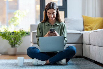 Wall Mural - Pretty young woman using her mobile phone while working with laptop sitting on the floor at home.