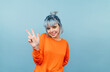 © bodnarphoto - Positive lady with blue hair standing on a blue background dressed in an orange sweater and showing a gesture of peace with a smile on her face.