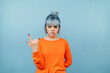 © bodnarphoto - Beautiful girl with blue hair and orange sweater stands on a colored background, bites her lip and shows a gesture of heavy metal to the camera