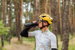 Handsome young man cyclist in helmet and goggles drinks water from a bottle on a background of pine forest and looks away.