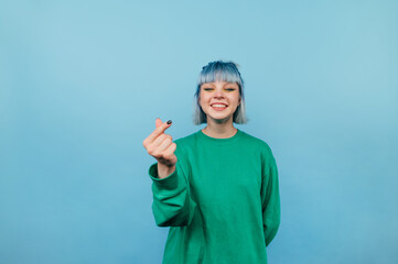 Positive hipster girl with colored hair shows heart gesture with fingers and looks at camera with smile on face on blue background.