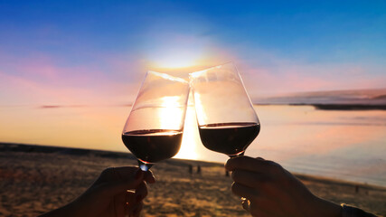 Wall Mural - Close up of couple hands with red of glass wine in a holiday with wine glasses of couple in a beautiful sunset on beach  background