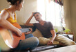 © Anne B/peopleimages.com - Does she know how amazing she is to me. Shot of a young woman playing the guitar while relaxing on the sofa with her husband.
