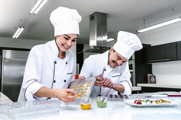 Wall Mural - young hispanic couple woman and man chocolatier in chef uniform and hat preparing mexican chocolates bonbon candies at kitchen in Mexico Latin America	