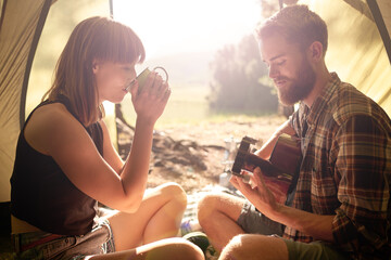 Wall Mural - Coffee time at camp. Shot of a young man playing guitar to his girlfriend in a tent.