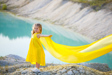 A small 6-7 years old Ukrainian girl in a yellow dress with wings in yellow fabric by the lake. Blue sky and yellow fabric, the colors of the Ukrainian flag.