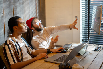 Two men with different nationality working on computers, sitting together at cozy home office. Concept of freelance and remote work. Stylish male hipsters programming together