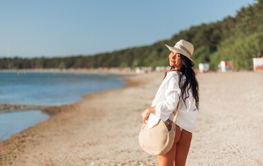 Wall Mural - people, summer holidays and leisure concept - happy young woman in white shirt and straw hat with bag walking along beach