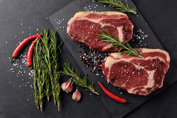 Raw meat, two beef steaks on a cutting board with rosemary, hot red pepper and spices on a black background, top view