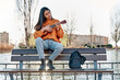 young latina playing ukulele in a city park. woman sitting on a bench practicing with her musical instrument.