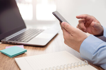 Businessman hold smartphone with finger touch to screen display and notebook and laptop on the table and working in office and glass window in blur background