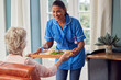 Female Care Worker In Uniform Bringing Meal On Tray To Senior Woman Sitting In Lounge At Home 