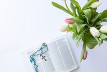 Open Christian Bible With Blue Rosary Prayer Beads Laying On A White Desk With A Vase Of Pink And White Tulips Shot From Above 