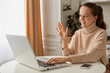 © sementsova321 - Indoor shot of adorable smile female woman with dark hair wearing beige sweater sitting in kitchen and working on laptop, having online conversation, video call.