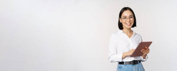 Wall Mural - Image of young asian woman, company worker in glasses, smiling and holding digital tablet, standing over white background