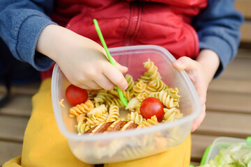 Little boy is eating his lunch after kindergarten or school from plastic container on bench in the park. Street take away food for child. Healthy meals for kids.