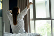 © thebigland45 - Young woman in a white t-shirt on the bed, stretching near the bed after waking up with sunrise in the morning. Beautiful woman looking outside her window.