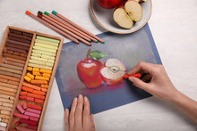 Woman drawing apples on paper with soft pastels at white table, top view