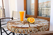 Orange juice in a tall transparent glass on an old metal table with beautiful patterns. Next to it is a fresh orange cut in half. A small metal table with chairs on the balcony.