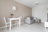 Fototapeta  - Przytulny salon w kolorach beżowych, białych i różowych. Szara sofa ze stolikiem na kawę. Stolik do jedzenia z wisząca różową lampą.