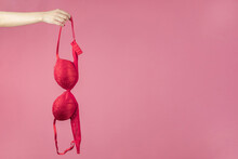 A Woman's Hand Holding A Red Bra On A Pink Background. The Concept Of Women's Breasts, Underwear. Copy Space