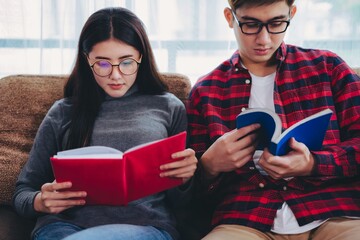 Two young adults sitting together on a couch, wearing glasses and focused on reading their books, creating a calm and studious atmosphere.