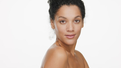 Close-up beauty portrait of attractive young slim African American woman with dark curly hair with bare shoulders looks at the camera over her shoulder on white background | Body care concept