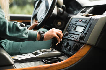 Caucasian female driver driving car, side view. Close-up of woman's hand holding an automatic transmission lever inside vehicle. Selective focus on female hand with watch