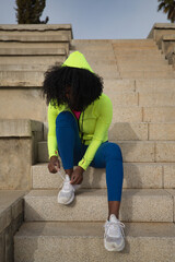 African-American woman with afro hair and sportswear, sitting, wearing a hood and leggings, buckling her laces to exercise in the street. Fitness concept, sport, street, urban.