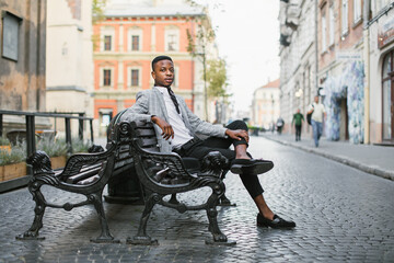Confident african businessman in stylish suit sitting alone on bench among city street. Handsome young guy enjoying free time on fresh air.