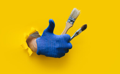 Wall Mural - The painter's hand in a blue knitted cloth glove holds two bristle brushes and shows a big stick up (like). Ripped hole in yellow paper, copy space. The concept of a worker, a labor migrant, a master.