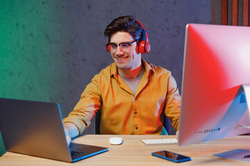 Young video content creator man in shirt headphones listen to music work at home sharing new production for his channel on laptop pc desktop computer, typing mounting content. Social media concept