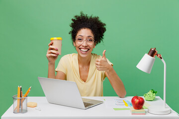 Wall Mural - Young employee business woman of African American ethnicity in shirt sit work at white office desk pc laptop hold takeaway delivery cup coffee to go show thumb up isolated on plain green background.