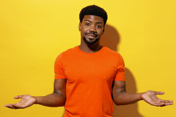 Wall Mural - Young man of African American ethnicity 20s in orange t-shirt spread hands shrugging shoulders look puzzled have no idea isolated on plain yellow background studio portrait People lifestyle concept