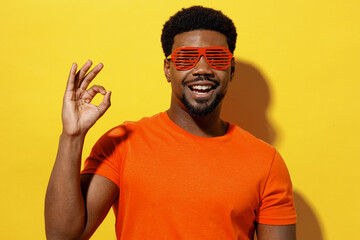 Wall Mural - Young smiling fashionable happy man of African American ethnicity 20s wear orange t-shirt stylish red glasses sho wokay gesture isolated on plain yellow background studio. People lifestyle concept