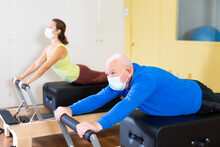 Mature Man In Protective Mask Practicing Pilates System On Reformer Supervised In Gym