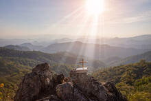 Crucifix Symbol And Bible On Top Mountain With Bright Sunbeam On The Colorful Sky Background