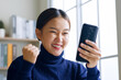 Young Asian woman using smartphone showing winner gesture, glad, excite at home