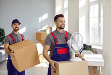 Male Porters From Moving Service Carry Cardboard Boxes With Things And Household Appliances. Two Smiling Handsome Professional Male Loaders In Overalls Help Client With Moving From Apartment Or Office