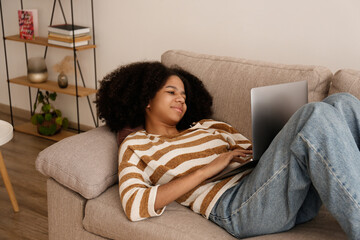 Black teenage girl with afro hairstyle lying on the couch, browsing the internet on her laptop. African american female wearing casual attire relaxing at home. Close up, copy space, background.