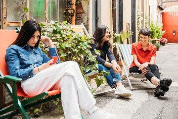 Three female co-workers on break relax outside the office on a sunny day sitting on deck chairs, one woman with smartphone while the others talk and joke - Millennials are resting near building