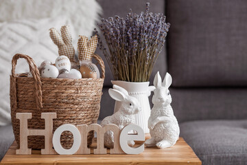 Wall Mural - Provence. A wicker basket with Easter eggs, lavender, candles and white rabbits in the interior of the living room on a wooden table. The concept of home comfort in the bright holiday of Easter 2022.