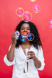 © beavera - Selective focus on soap bubble blown by unrecognizable woman in red background. Vertical front view of african american woman playing funny with soap bubbles. Party background concept.