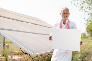 Smiling village farmer showing white empty board by looking at camera in front of solar panel at farmland - concept of promotion, advertisement and renewable energy.