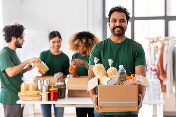 Wall Mural - charity, donation and volunteering concept - happy smiling male volunteer with food in box and international group of people at distribution or refugee assistance center