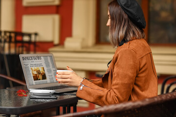 Sticker - Young woman checking news on laptop and holding cup of coffee in street cafe