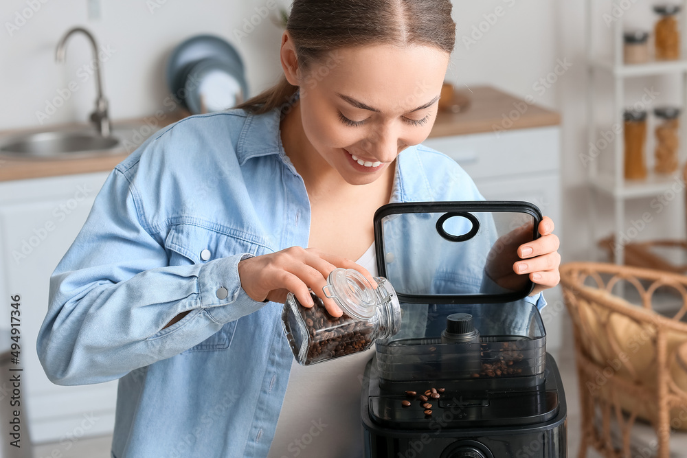 Young woman putting beans into coffee machine in kitchen, closeup