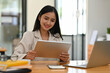 © saltdium - A portrait of a beautiful smiley Asian businesswoman sitting in the office holding a tablet and working on a laptop, for business and technology concept.