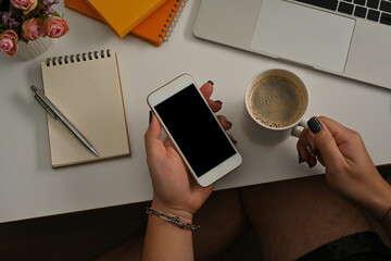 Poster - Top view of hand holding a smartphone and a cup of coffee leaning to the table have a pen, note books and laptop on the side, for business and technology concept.
